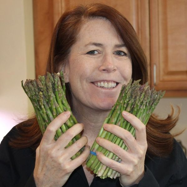 Woman smiling and holding two bunches of asparagus in a kitchen.