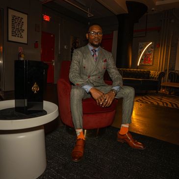 A stylish man in a gray suit with orange socks sits confidently on a red chair indoors.