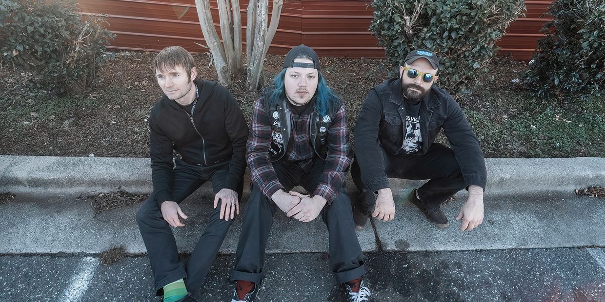 Three men sitting on a curb with trees and bushes behind.