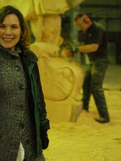 A woman smiles in the foreground, while a man carves wood in the background.