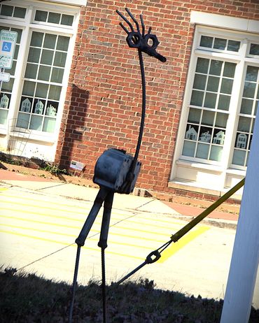 Metal sculpture of a whimsical bird standing outdoors near a brick building.