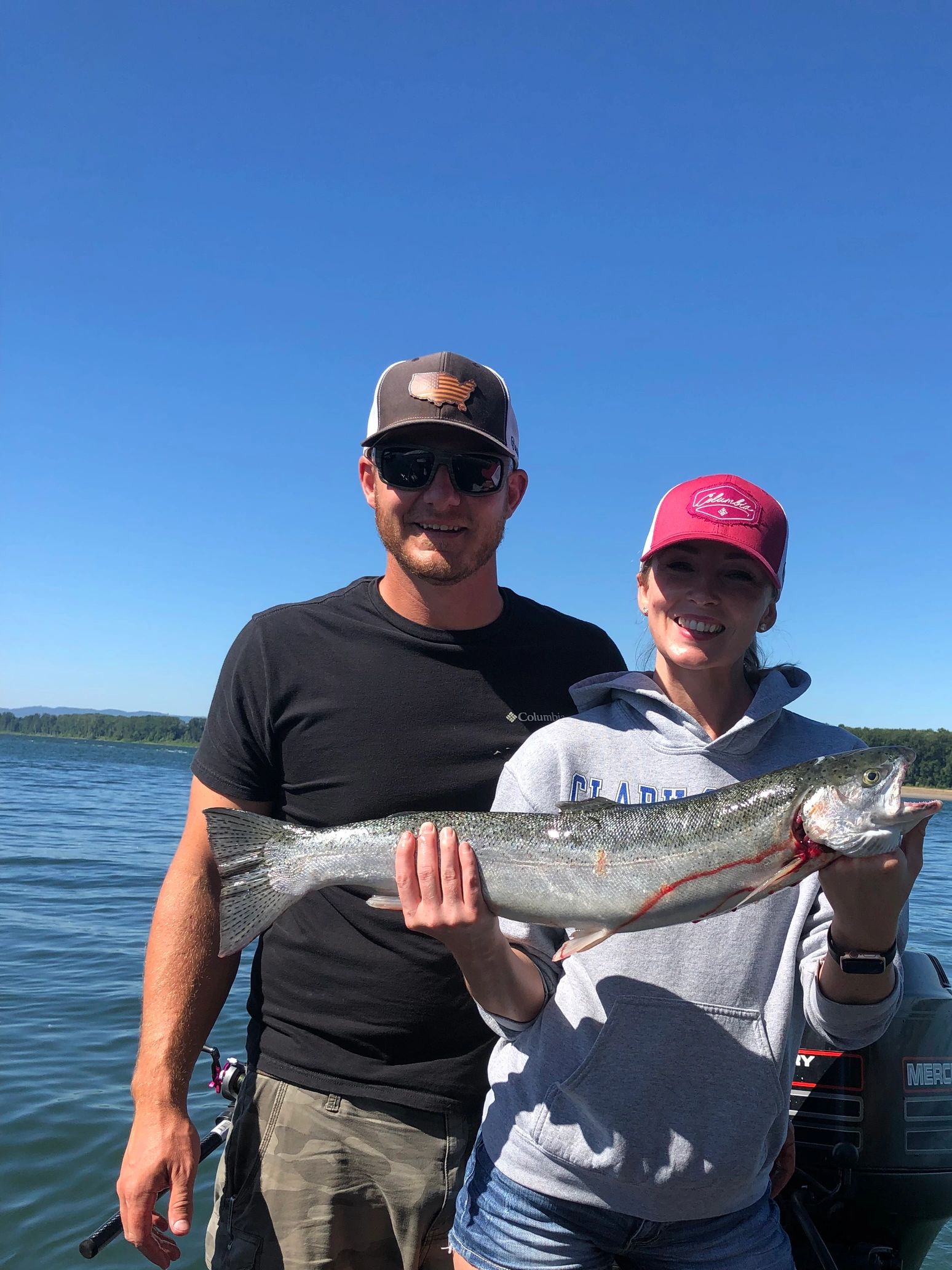 A smiling couple holding a large fish on a sunny day by the water.