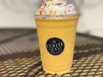 A creamy yellow smoothie topped with whipped cream and colorful sprinkles in a clear cup.