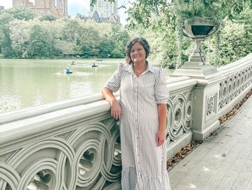 Woman in striped dress stands by ornate bridge railing with lake and cityscape in background.