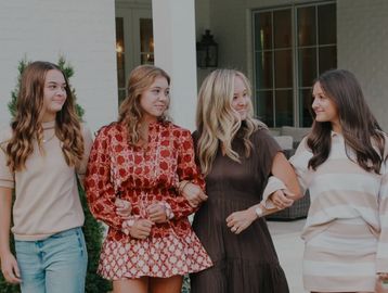 Four women walking arm in arm outside a house, smiling at each other.