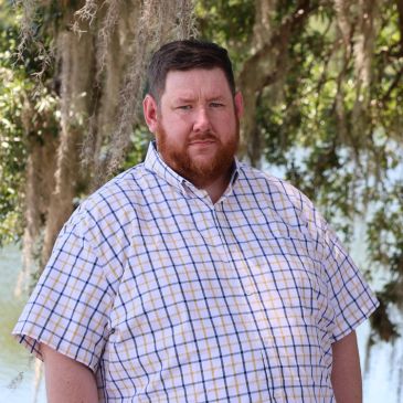 A man with a beard in a checkered shirt outdoors near trees.