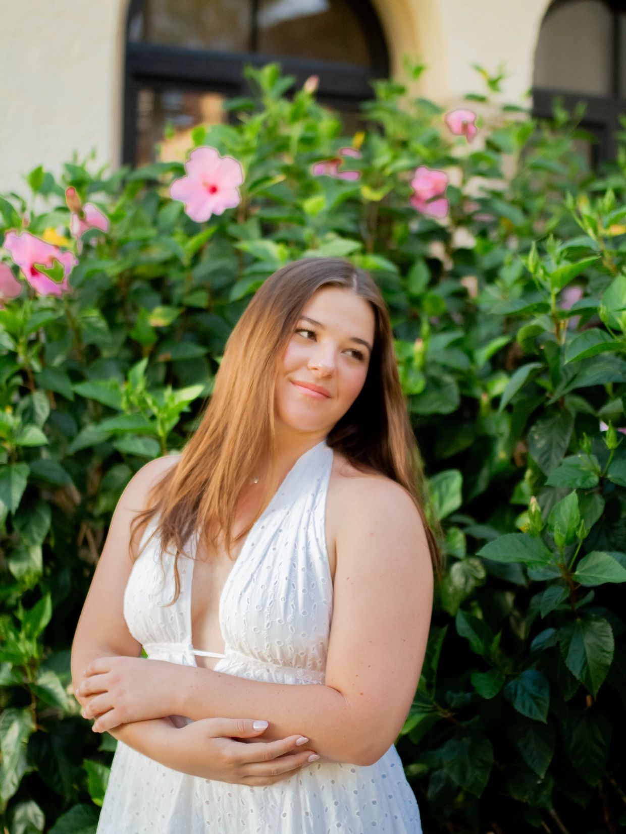 Young woman in white dress posing by pink flowers and green leaves.