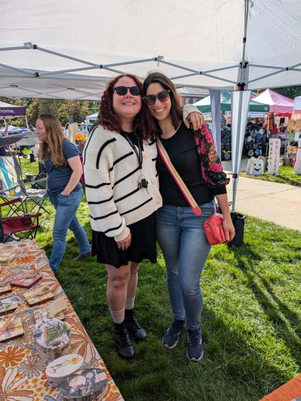 Two women smiling outdoors at a market or fair.