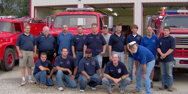 Group of firefighters posing in front of fire trucks outside a fire station.