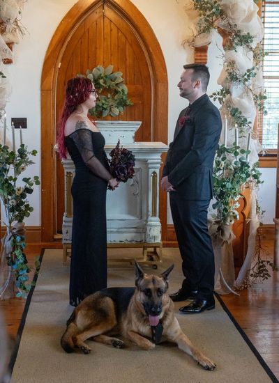 Couple in black attire exchanging vows with a German Shepherd lying in front.