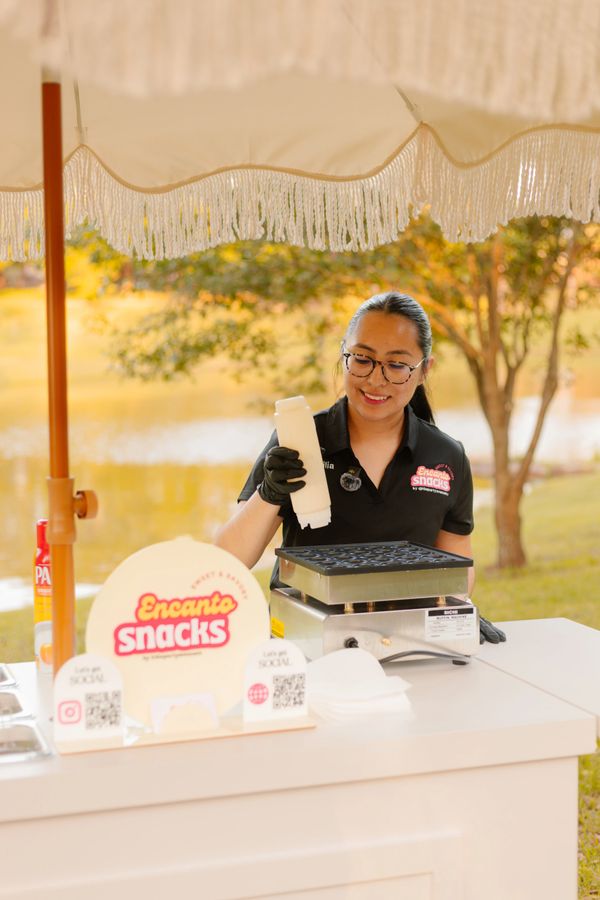 Woman preparing snacks at an outdoor food stall under an umbrella.