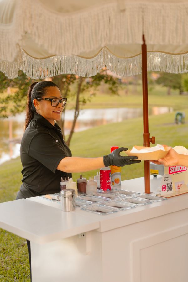 Woman serving food from an outdoor food cart under an umbrella.