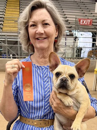 Woman proudly holding a ribbon and her French Bulldog at a competition.