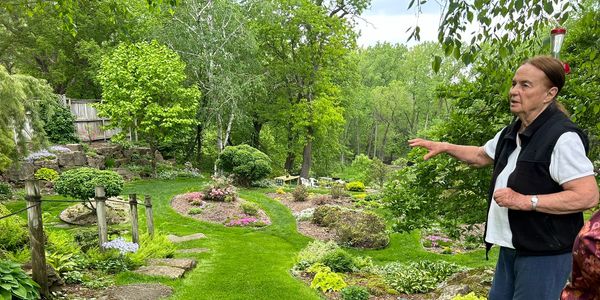 An elderly woman gestures while explaining a lush garden with blooming flowers and greenery.