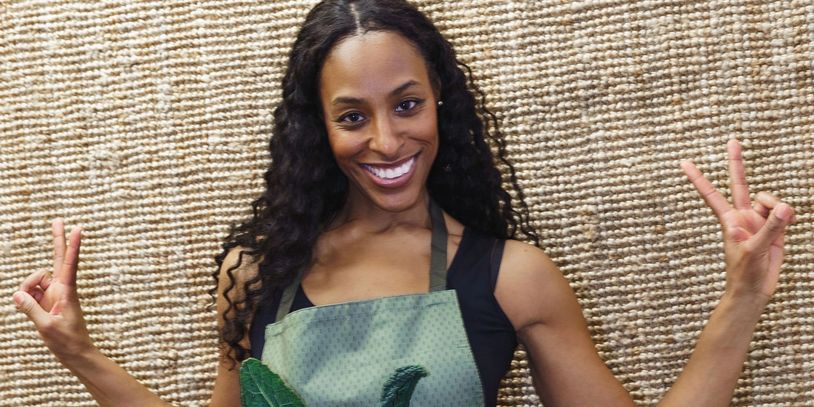 Smiling woman in apron holding vegetables and making peace signs.