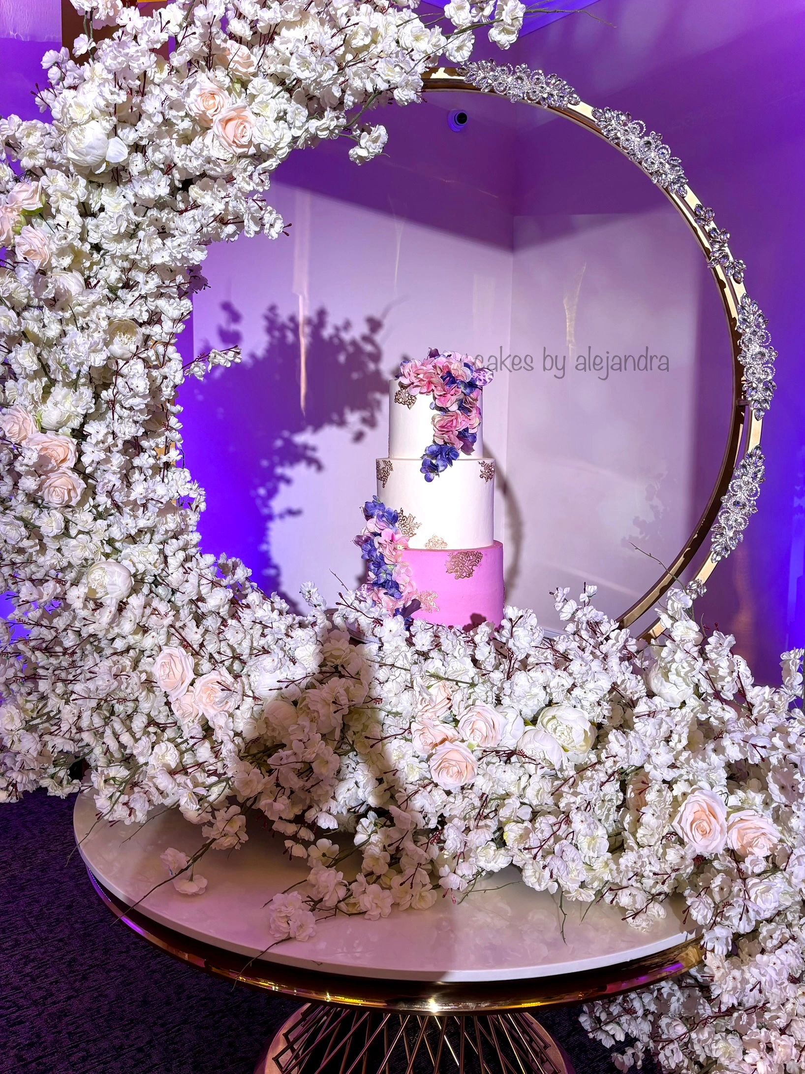 Elegant three-tiered cake surrounded by white flowers and a jeweled circular frame.