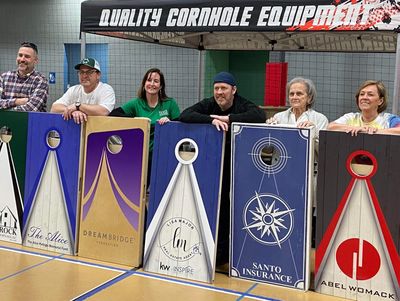 Six people stand behind customized cornhole boards under a Quality Cornhole Equipment tent.