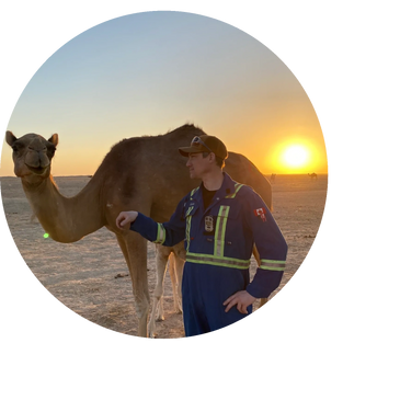 A man in workwear stands beside a camel at sunset in a desert.