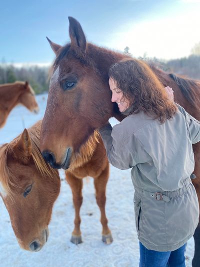 A woman affectionately hugs two horses in a snowy outdoor setting.