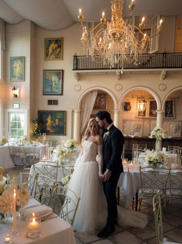 Bride and groom holding hands in an elegant wedding venue with chandeliers and floral decorations.