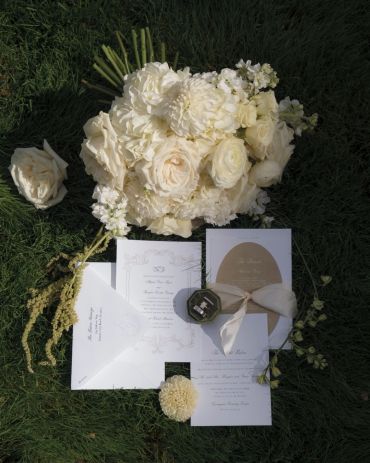 Elegant white floral bouquet with wedding invitations on grass.