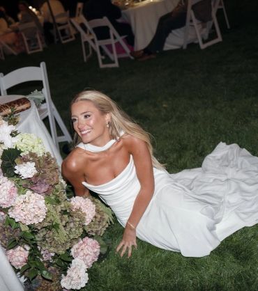 Bride in white dress sitting on grass near flowers, smiling.