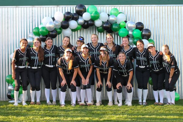 A girls' softball team poses together under a colorful balloon arch.