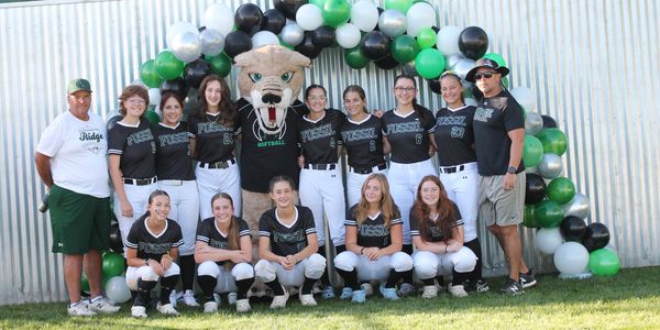 Softball team posing with mascot and coaches under balloon arch.