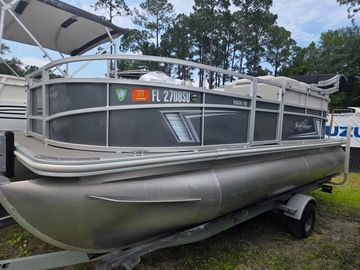 Gray pontoon boat on trailer near trees.