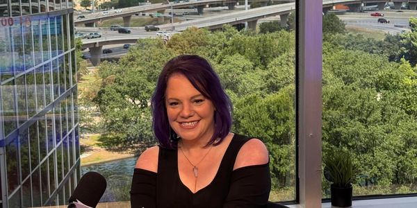 Woman with purple hair smiling in a studio with a city view behind her.