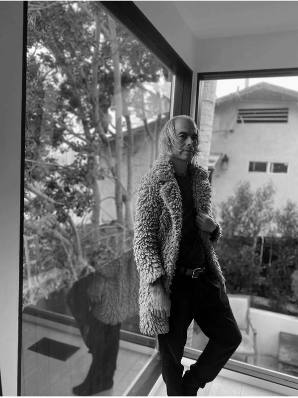 A man in a textured coat stands by a large window, looking contemplative.