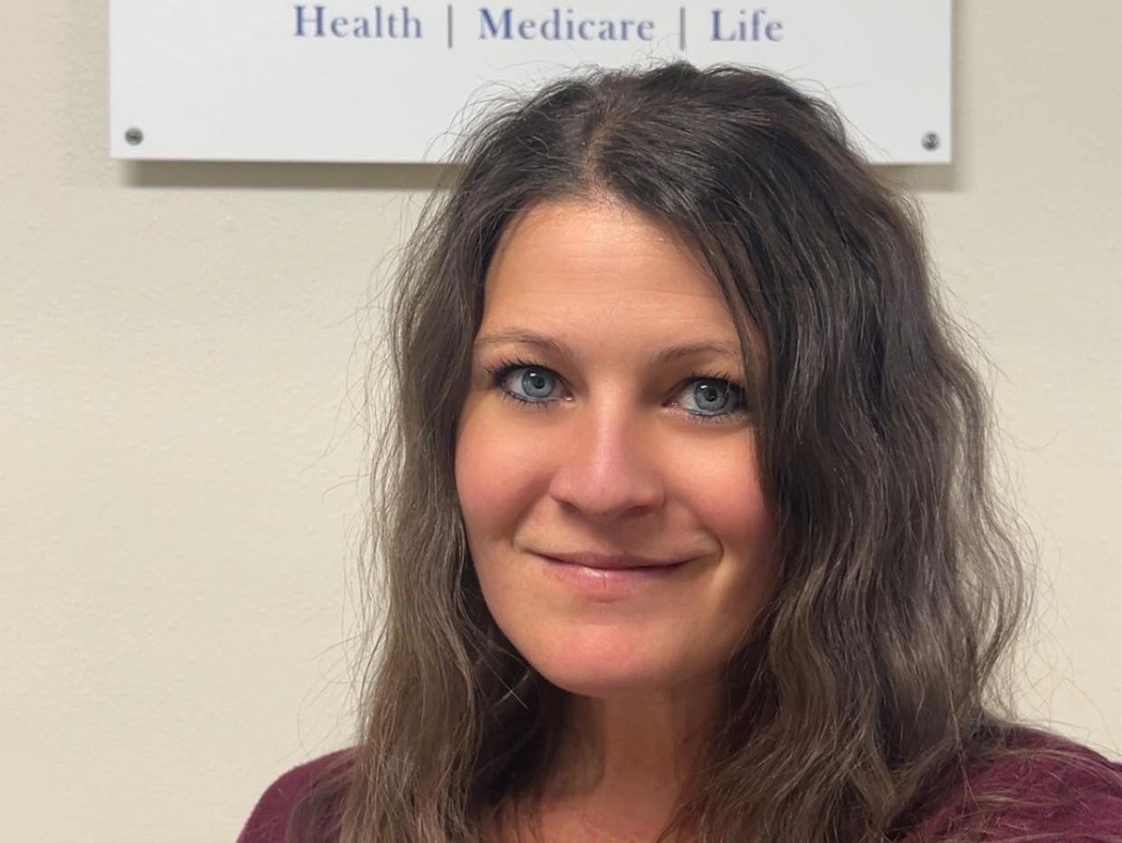 Smiling woman with blue eyes and wavy brown hair in front of a healthcare sign.