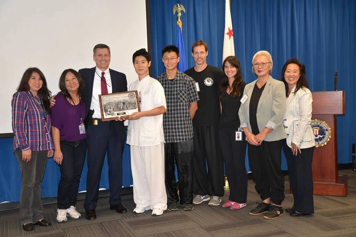 A diverse group poses with an award plaque in a formal setting with flags.