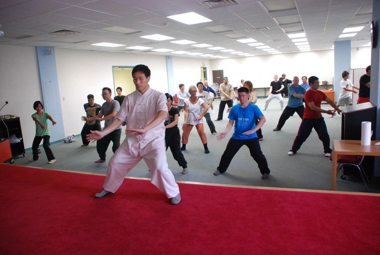 A group practicing Tai Chi in a spacious indoor room led by an instructor.