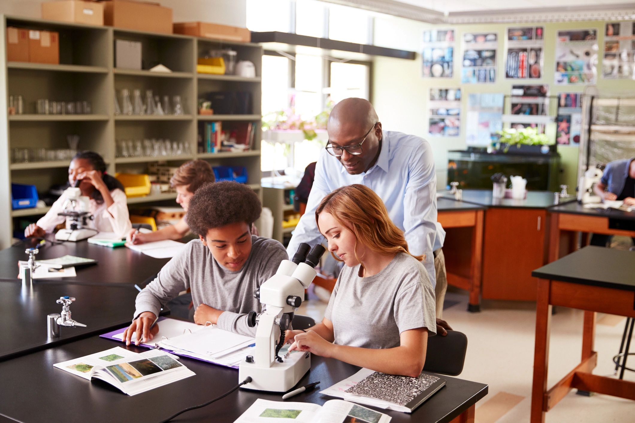 Teacher looking over the shoulders of two students working on their assignment with a stereo microsc