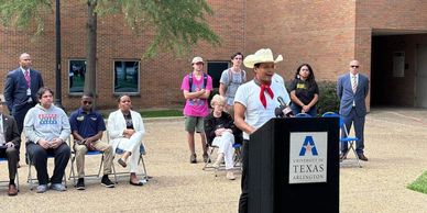 Man in cowboy hat speaking at a University of Texas Arlington event outdoors.