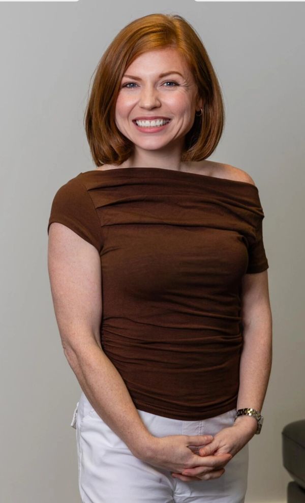 Smiling woman with red hair in brown top and white pants.