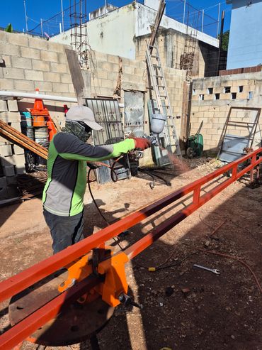 Worker spray painting a long metal beam bright orange outdoors.