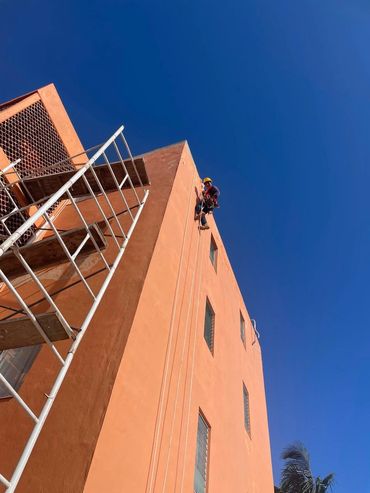 Worker descends orange building with safety gear against clear blue sky.