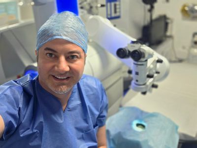 Surgeon in blue scrubs smiling in an operating room with medical equipment.