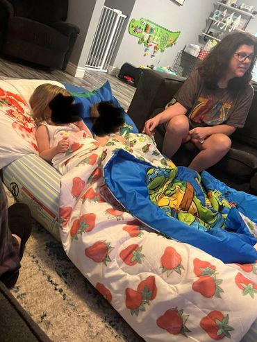 Two children lying under blankets and a woman sitting on a couch in a living room.