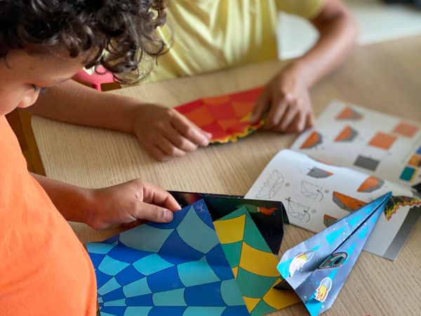 Two children folding colorful paper planes with instructions on the table.