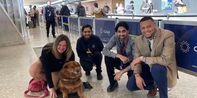 Four people smiling with a fluffy dog at an indoor public space.