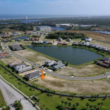 Aerial view of a suburban neighborhood with a pond and empty plot marked by an orange arrow.