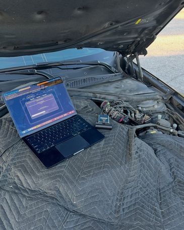 A laptop and diagnostic tool set up on a padded cover inside a car engine bay.