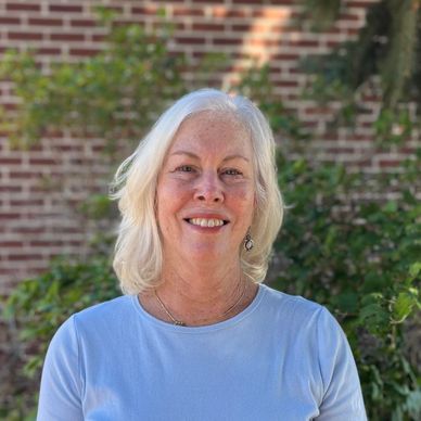 Smiling woman with white hair outdoors against a brick and greenery background.