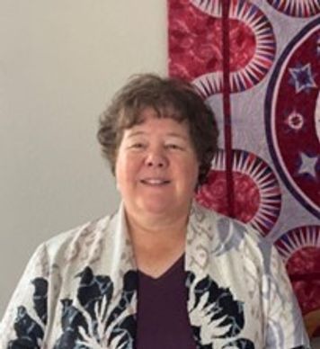 Smiling woman with short curly hair standing indoors near a colorful tapestry.