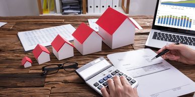 Person calculating finances with house models and laptop on desk.