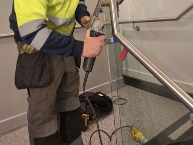 Worker using a power tool on a glass panel near a staircase.