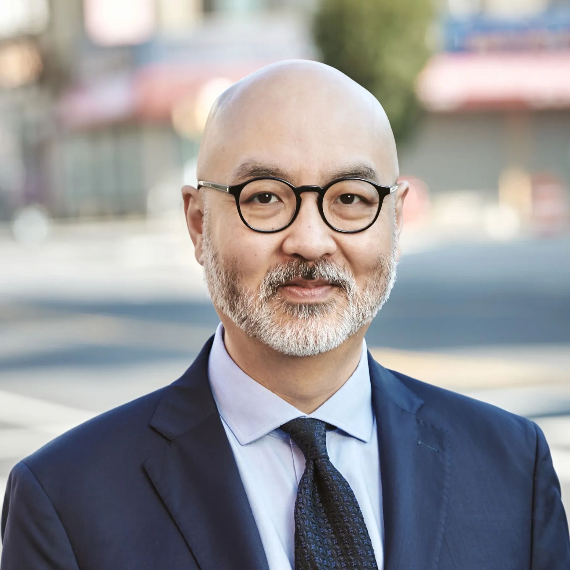 Smiling bald man with glasses wearing a navy suit and tie outdoors.